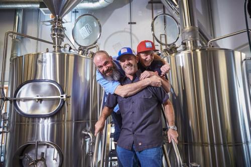 Three men smile and pose playfully in a brewery, surrounded by large brewing equipment.