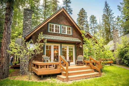 Cozy wooden cabin surrounded by trees, featuring a deck with chairs and a stone chimney.