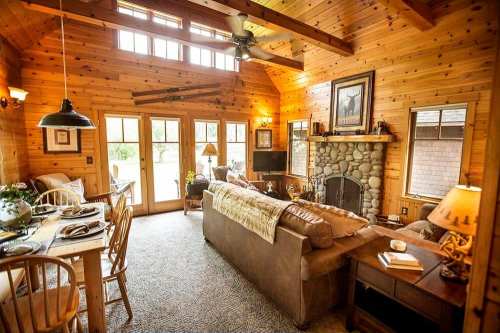 Cozy wooden cabin interior with a stone fireplace, comfortable seating, and a dining area, illuminated by warm lighting.