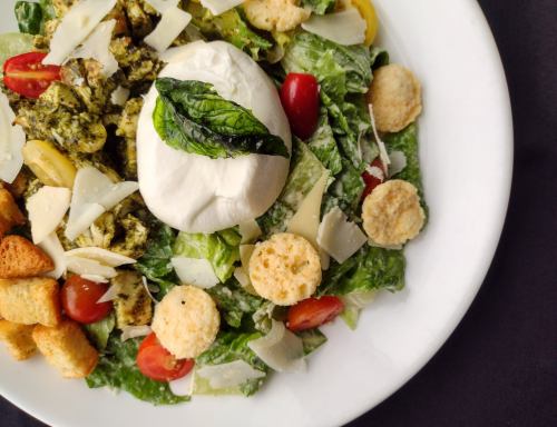 A fresh salad with romaine, cherry tomatoes, croutons, parmesan, pesto, and a creamy burrata topped with basil.