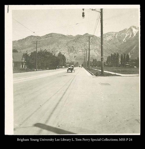 A vintage photo of a wide road lined with trees and houses, with mountains in the background.
