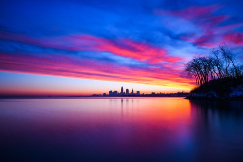 Vibrant sunset over a city skyline, reflecting colorful clouds on calm water, with silhouetted trees in the foreground.