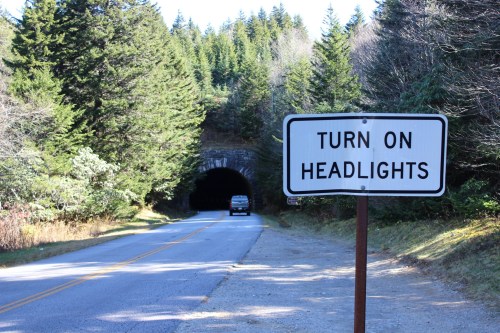 A road sign reading "Turn on headlights" near a tunnel entrance, surrounded by trees and a vehicle approaching.
