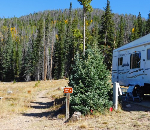 A camper parked near a trail sign in a forested area with tall trees and autumn foliage.