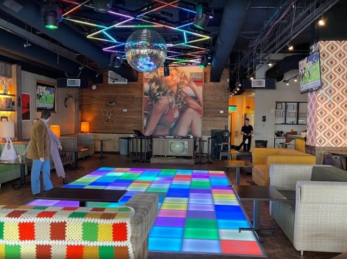 A vibrant lounge with a colorful dance floor, disco ball, and retro decor, featuring a large wall mural and cozy seating.