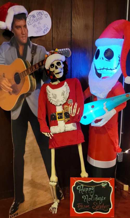 A skeleton in a Santa shirt stands next to a cardboard Elvis and a festive skeleton holding a guitar, all decorated for the holidays.