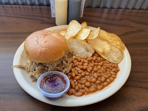 A plate with a pulled pork sandwich, baked beans, and potato chips, served with a side of barbecue sauce.