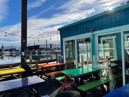 Colorful picnic tables on a deck overlooking a marina with boats and a blue sky.