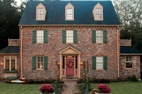 A charming brick house with green shutters, a pink door, and a welcoming front garden adorned with flowers and pumpkins.