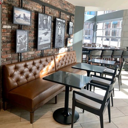 Cozy café interior with a brown tufted bench, glass tables, and framed photos on a brick wall. Bright, modern atmosphere.
