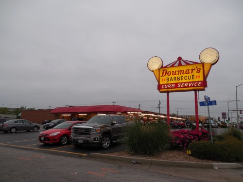 Sign for Doumar's Barbecue with curb service, featuring a red building and parked cars under a cloudy sky.