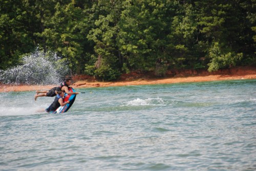 Two people ride a colorful inflatable on a lake, splashing water as they speed across the surface. Green trees in the background.