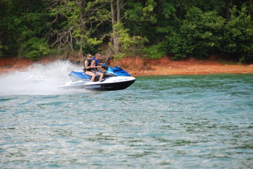 Two people ride a blue jet ski across a lake, creating a spray of water against a backdrop of green trees.