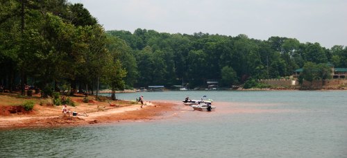 A serene lakeside scene with boats anchored near a sandy shore, surrounded by lush green trees.