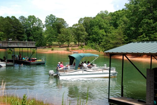 A pontoon boat with passengers navigates a calm lake surrounded by green trees and a dock in the background.
