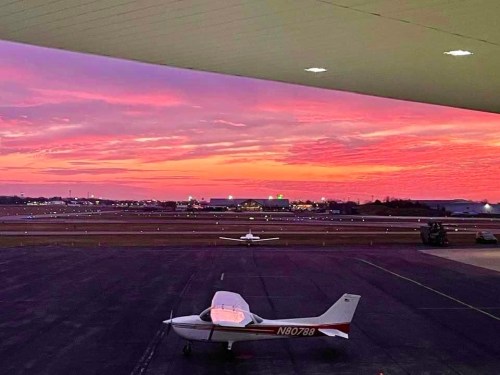A small airplane on the tarmac with a vibrant sunset sky in the background, casting colorful hues over the scene.