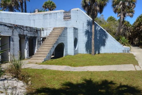 A blue concrete structure with stairs, surrounded by grass and palm trees under a clear blue sky.