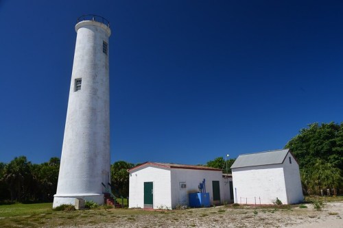 A tall white lighthouse beside a small white building, set against a clear blue sky and green foliage.