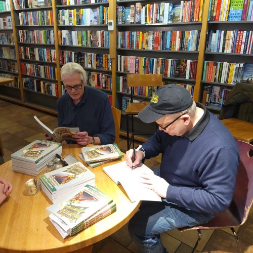 Two men sit at a table in a bookstore, one reading and the other signing a book, surrounded by shelves of books.