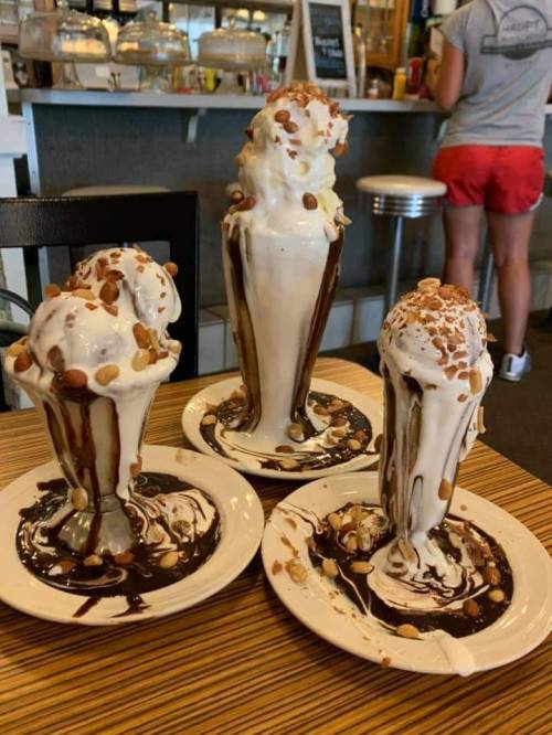 Three overflowing ice cream sundaes with whipped cream, nuts, and chocolate sauce, on plates with melted ice cream.