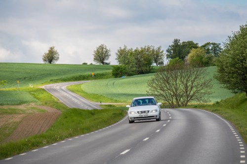 A silver car drives along a winding road surrounded by green fields and trees under a cloudy sky.