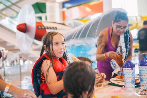 A girl in a life jacket enjoys food at a water park, with a colorful slide and a woman nearby.