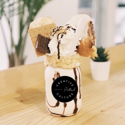 A decadent milkshake topped with whipped cream, chocolate drizzle, graham crackers, and a slice of cake in a jar.