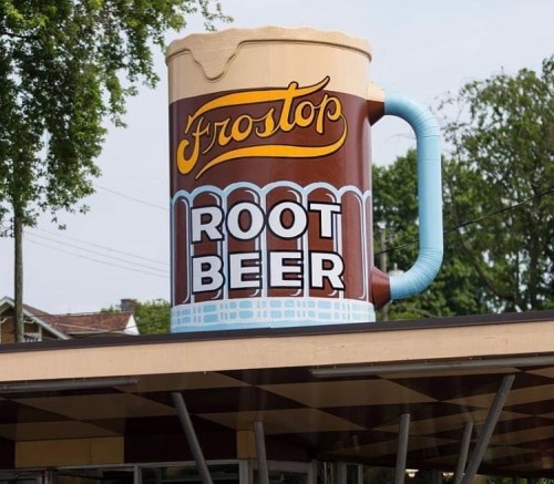 A large, colorful sign of a frosty root beer mug with "Frostop Root Beer" written on it, set against a tree-lined background.