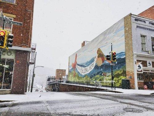 A snowy street scene featuring a colorful mural on a building that reads "You Belong Here." Traffic lights visible.