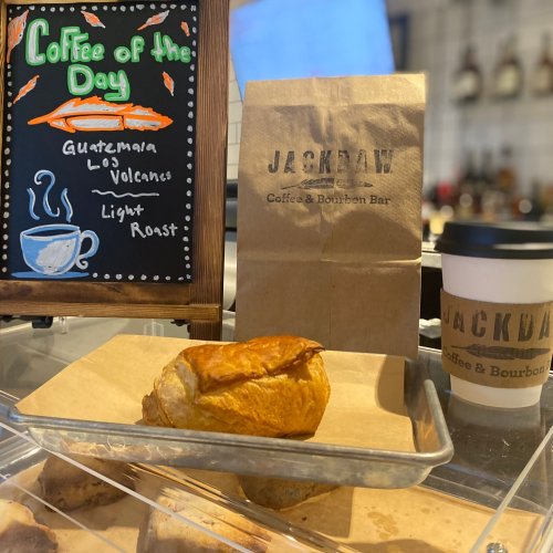 A chalkboard displays "Coffee of the Day" with a light roast. A pastry sits on a tray next to a coffee cup and a bag.