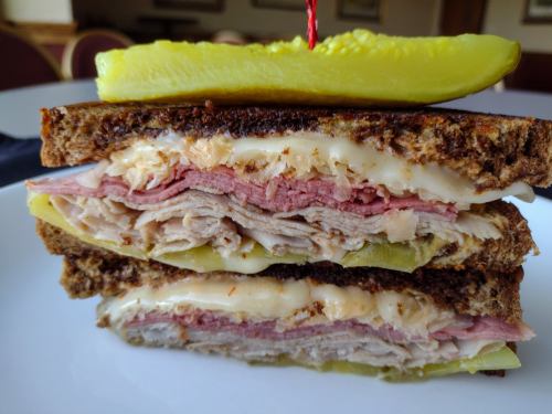 A stacked sandwich with layers of turkey, ham, and pickles, served on dark bread with a pickle on top.