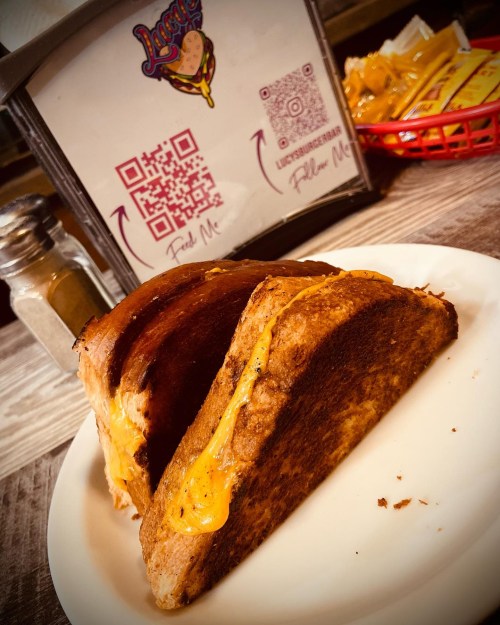 A grilled cheese sandwich on a plate, with a QR code sign and a basket of fries in the background.
