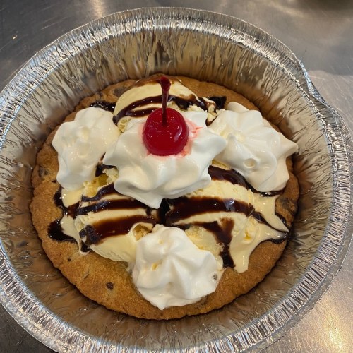 A dessert featuring a large cookie topped with ice cream, whipped cream, chocolate sauce, and a cherry.