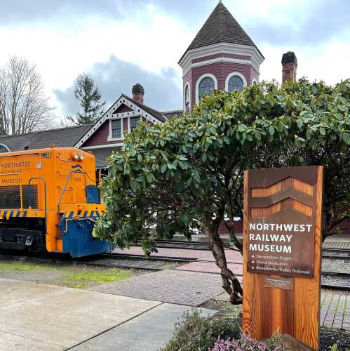 A bright orange train near a historic building, with a sign for the Northwest Railway Museum in a scenic setting.