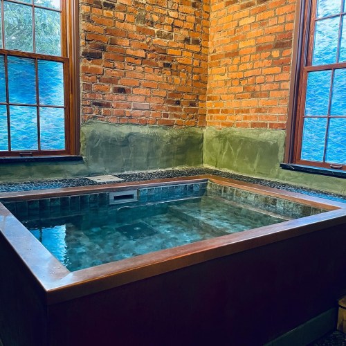 A small indoor hot tub surrounded by brick walls and large windows, with water reflecting the light.