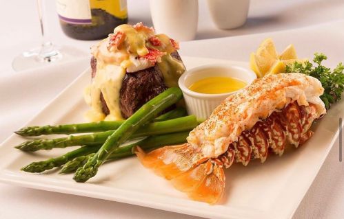 A gourmet plate featuring filet mignon topped with lobster, asparagus, and a side of lemon and butter sauce.