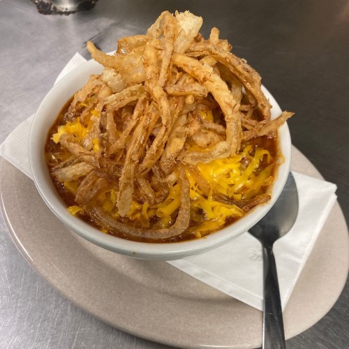 A bowl of chili topped with shredded cheese and crispy fried onions, served on a plate with a spoon.