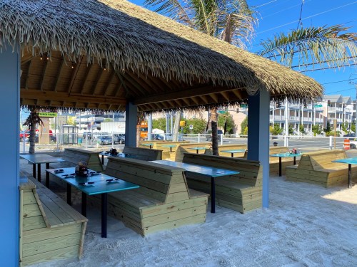 A beachside dining area with wooden benches and a thatched roof, surrounded by palm trees and sandy ground.