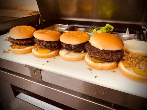 Five burger patties on buns, with pickles and onions, lined up on a cutting board in a kitchen setting.