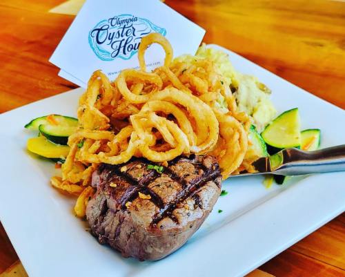 A plate featuring a grilled steak, curly fries, and zucchini, with a card for Olympia Oyster House in the background.