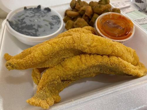 A takeout container with three fried fish fillets, a side of okra, a cup of sauce, and a serving of greens.