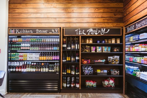 A well-organized pantry display featuring beverages and snacks on wooden shelves with chalkboard labels.
