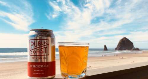 A can of Public Coast 67 Blonde Ale next to a glass of beer, with a beach and rock formation in the background.