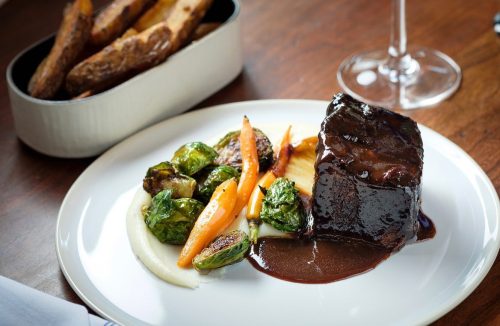 A plated dish featuring braised meat, roasted vegetables, and creamy puree, with a side of fries and a glass of wine.