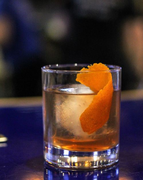 A glass of cocktail with a large ice cube and an orange peel garnish, set on a blue surface.