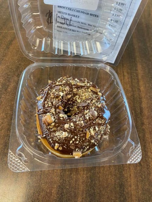 A chocolate-covered donut topped with crushed nuts, displayed in a clear plastic container.