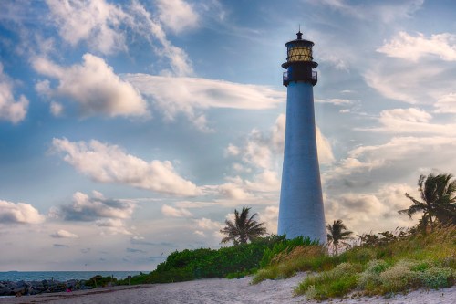 A tall lighthouse stands against a cloudy sky, surrounded by greenery and sandy beach, with the ocean in the background.