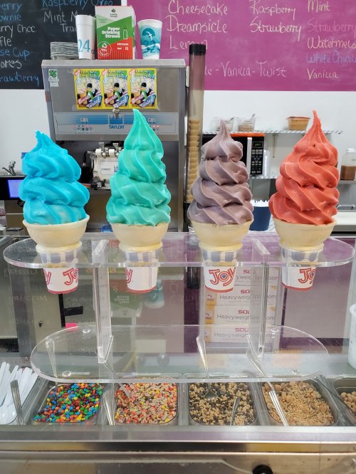 Four colorful soft serve ice cream cones in blue, teal, chocolate, and orange, displayed in a shop.
