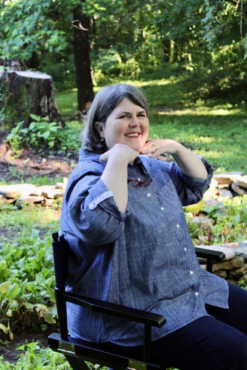 A woman with gray hair sits in a chair outdoors, smiling and playfully posing with her hands near her shoulders.
