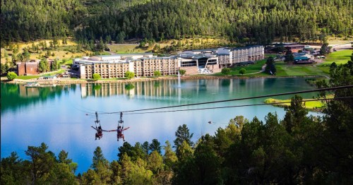 A scenic view of a lake surrounded by trees, with a hotel and zip line in the foreground.